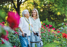 An adult granddaguhter supporting her senior grandmother when taking her for walk with walker in park in summer.
